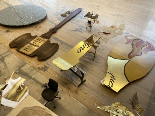 Überdimensionale Requisiten, darunter ein Richterhammer mit "Justice" und goldene Schriftzüge, liegen auf einem Werkstattboden.