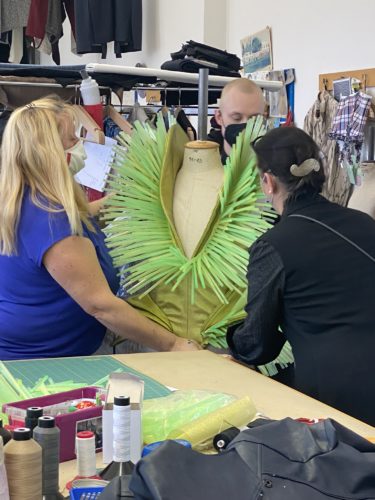 Drei Personen arbeiten gemeinsam an einem ausgefallenen grünen Kostüm mit stacheligen Verzierungen in einem Schneideratelier.