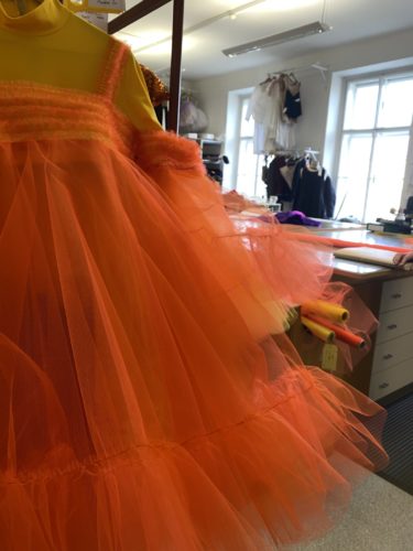 Ein leuchtend orangefarbenes Tüllkleid hängt in einer Schneiderei neben Arbeitsflächen und Nähmaterialien.