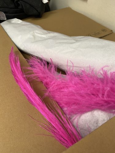 Leuchtend pinke Federn liegen auf braunem Karton, daneben eingewickeltes weißes Papier.