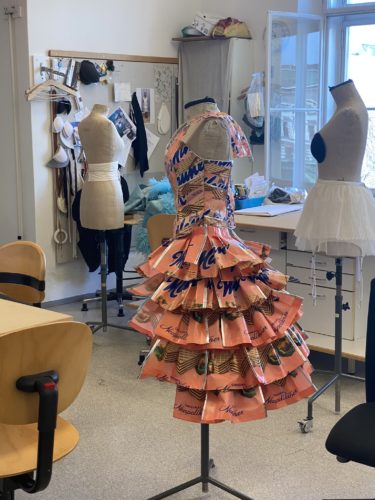 Ein Schaufensterpuppe trägt ein auffälliges Kleid aus bedrucktem Papier in einem hellen Atelier mit weiteren Schneiderpuppen und Nähmaterial.