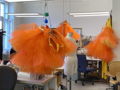 Mehrere orangefarbene Tutu-Kleider hängen in einem hellen Schneideratelier neben Schneiderpuppen und Nähmaschinen.