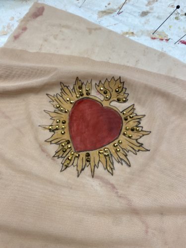 Ein rotes Herz, umgeben von goldener, flammenartiger Verzierung und kleinen goldenen Perlen, auf beigem Stoff gemalt.