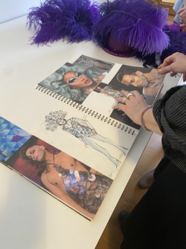 Jemand blättert in einem Skizzenbuch mit Zeichnungen und Fotos von Personen in farbenfrohen, aufwendigen Kostümen und Make-up.