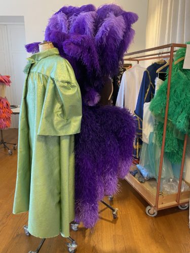 Ein glänzender, grüner Mantel und ein opulenter Kopfschmuck mit lila Federn hängen auf Kleiderständern in einem Ankleidezimmer.