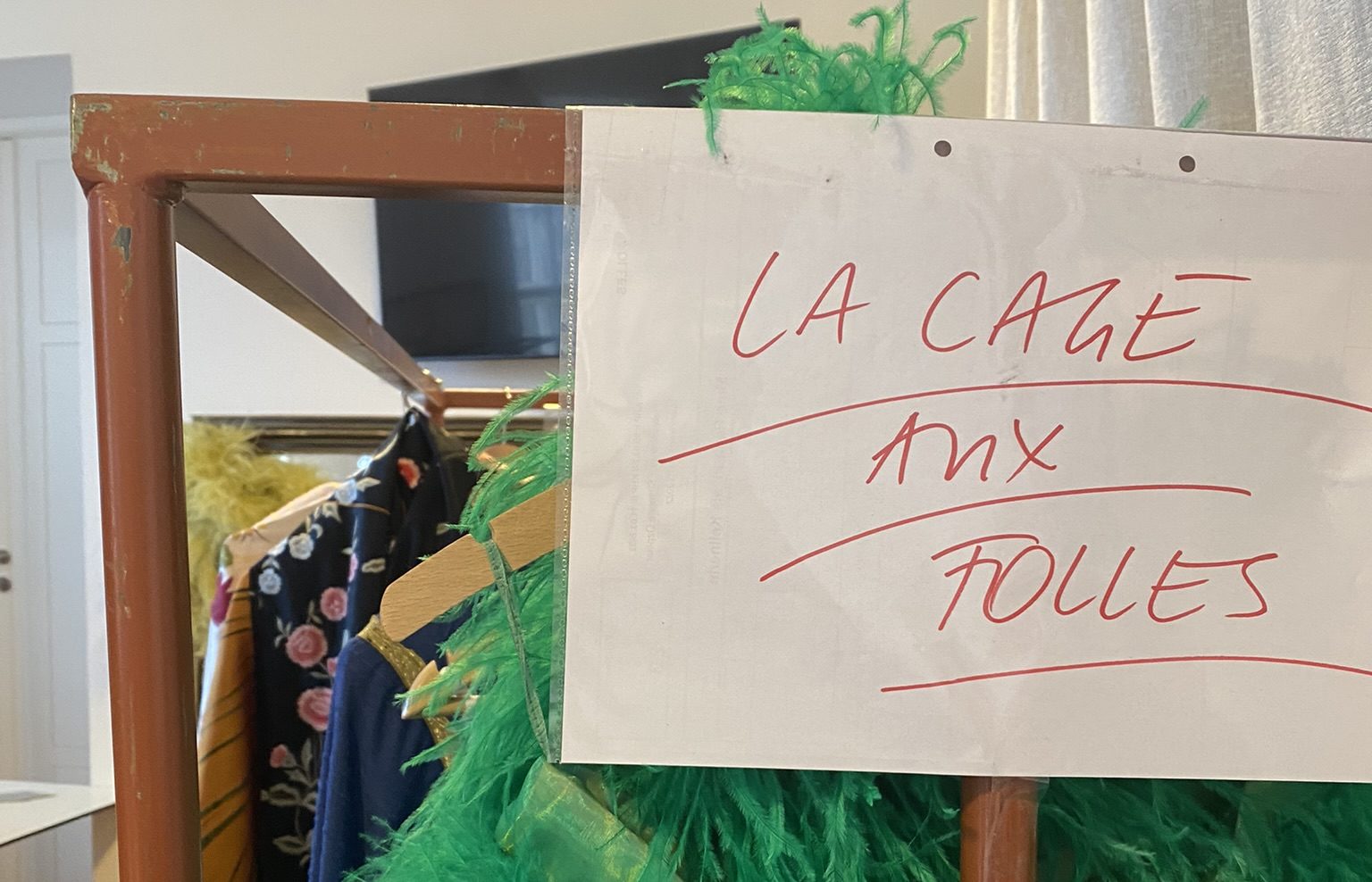 Kleiderständer mit bunten Kostümen, daran ein handgeschriebenes Schild mit der Aufschrift „La Cage aux Folles“.