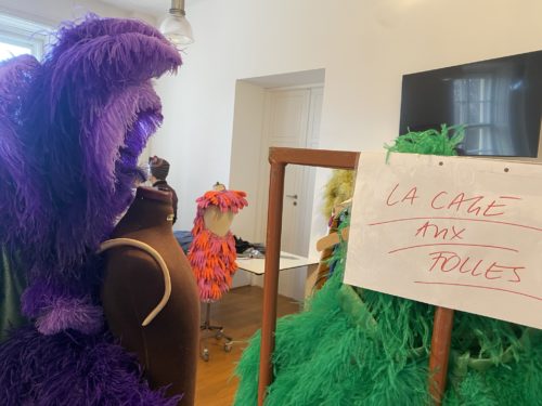 Eine Schneiderpuppe trägt ein lila Federkostüm, daneben hängen weitere farbenfrohe Kostüme und ein Schild mit der Aufschrift „La Cage aux Folles“.