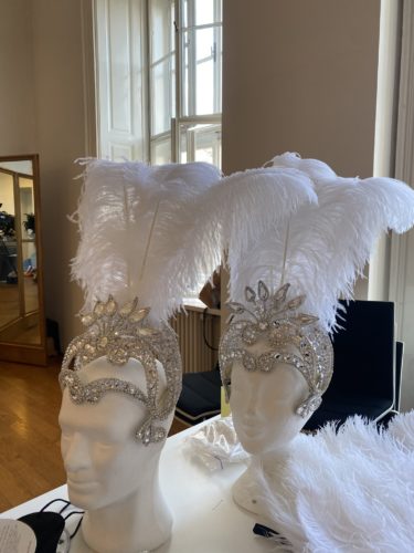 Zwei weiße Schaufensterpuppen mit glitzernden, silbernen Kopfbedeckungen und großen weißen Federbüscheln stehen auf einem Tisch.