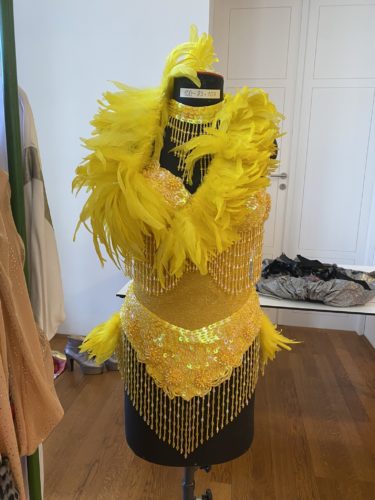 Ein gelbes Paillettenkostüm mit großen gelben Federn und funkelnden Fransen hängt auf einer Schneiderpuppe in einem Zimmer.