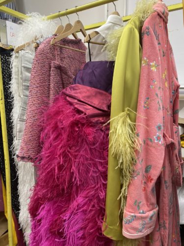 Mehrere bunte Kleidungsstücke hängen auf einer Stange, darunter ein pinkes Federkleid, eine gelbe Jacke mit Federn und ein rosa Bademantel mit Blumenmuster.