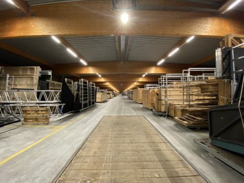 Lagerhalle mit Regalen und gestapelten Holzplatten sowie Metallgestellen entlang eines langen Mittelgangs.