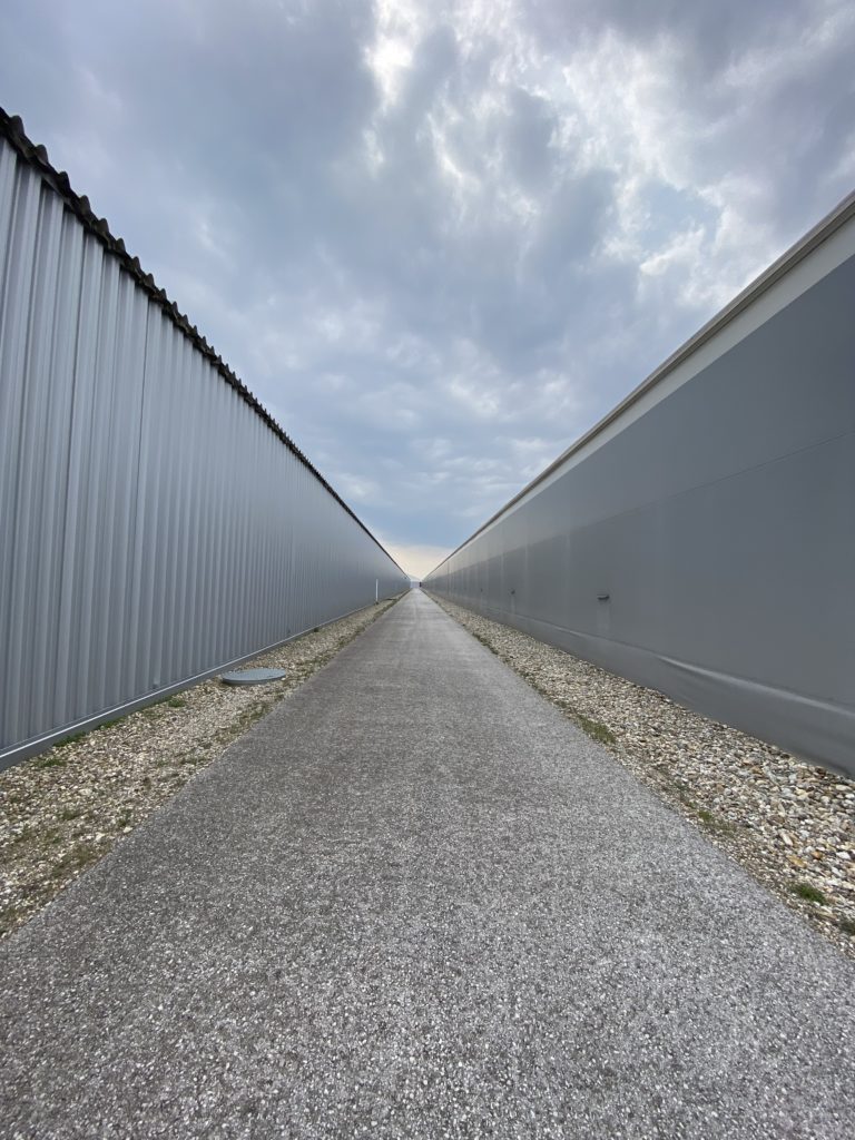 Langer, gerader Weg zwischen zwei grauen Industriegebäuden unter bewölktem Himmel.