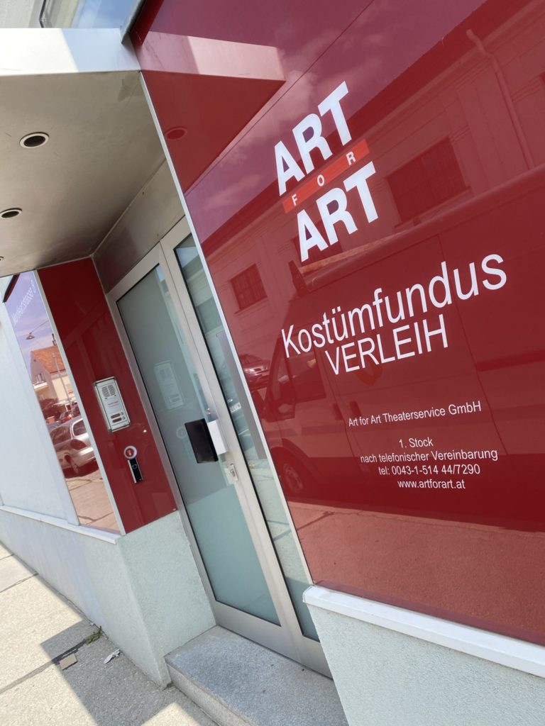 Roter Eingang mit Glastür und weißen Schriftzügen „ART FOR ART“ und „Kostümfundus VERLEIH“ an der Fassade.
