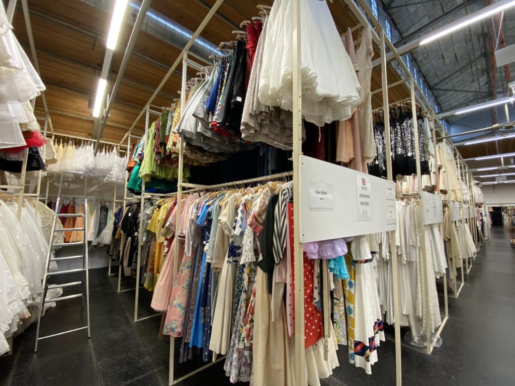 Viele bunte Kleider hängen ordentlich auf Kleiderstangen in einer großen Halle mit hell erleuchteter Decke.