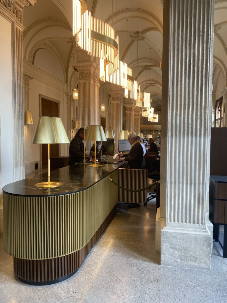 Mehrere Personen arbeiten an Schreibtischen mit goldenen Lampen in einer eleganten Halle mit hohen Decken und Säulen.