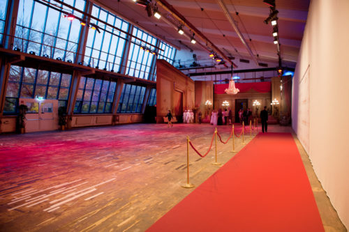 Großer Festsaal mit rotem Teppich, goldener Absperrung und elegant gekleideten Gästen in der Ferne, Kronleuchter und Bühnenbeleuchtung.