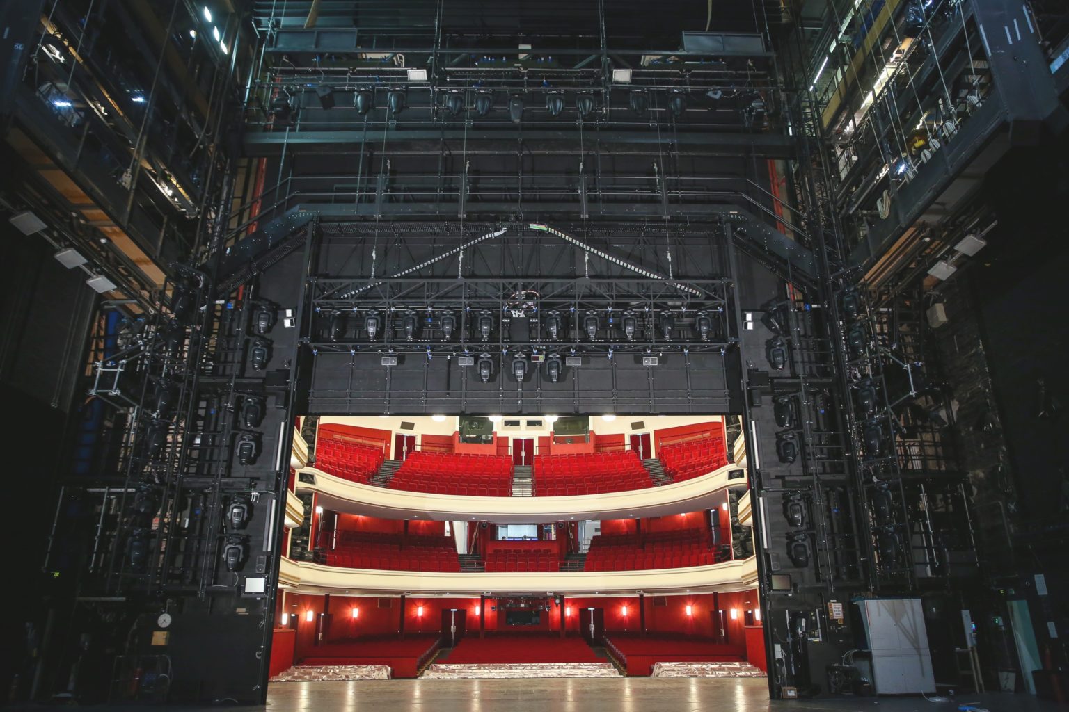Blick von der Bühne in einen leeren Theatersaal mit roten Sitzreihen und zwei Rängen.