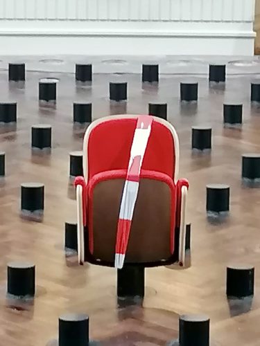 Ein einzelner roter Stuhl mit rot-weißem Absperrband steht auf einem Holzboden, umgeben von vielen schwarzen Zylindern.