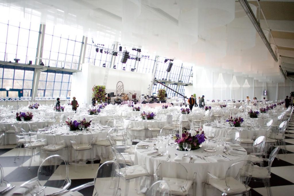 Eleganter Bankettsaal mit runden, weiß gedeckten Tischen und lila Blumenarrangements, große Fenster im Hintergrund.