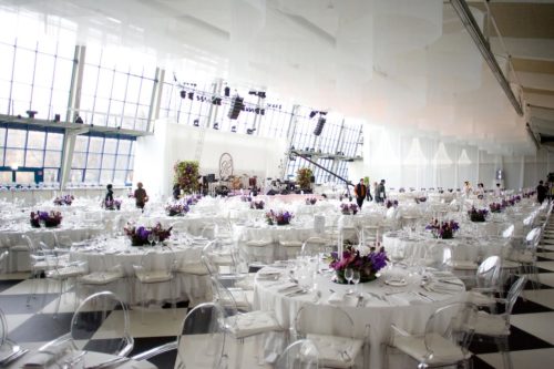 Eleganter Bankettsaal mit runden, weiß gedeckten Tischen und lila Blumenarrangements, große Fenster im Hintergrund.