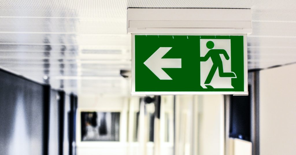 Grünes Notausgangsschild mit weißem Pfeil nach links und laufender Person an einer Decke in einem Flur.