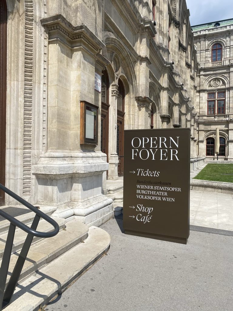 Großes braunes Schild vor einem historischen Gebäude mit der Aufschrift „OPERN FOYER“, das auf Tickets, Shop und Café hinweist.