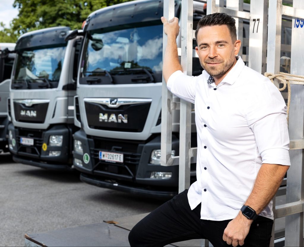 Ein Mann in einem weißen Hemd steht vor mehreren MAN-Lkw und lächelt in die Kamera.