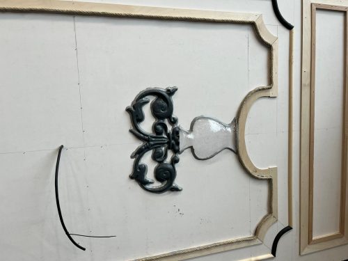 Verzierte Wandverkleidung mit aufwendigen Stuckelementen und geschwungenen Linien auf hellem Untergrund.