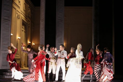 Eine Gruppe von Darstellern in historischen Kostümen steht auf einer Theaterbühne vor hohen Fensterläden, manche in roten Uniformen, andere in Kleidern.