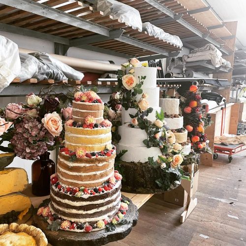 Mehrstöckige Hochzeitstorten mit Blumen, Beeren und Obst dekoriert stehen nebeneinander in einer Werkstatt mit Regalen und Stoffrollen.