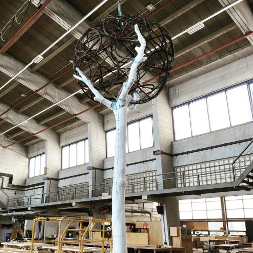 Ein weiß gestrichener künstlicher Baum mit einer kugelförmigen Skulptur aus Ästen steht in einer Werkhalle.