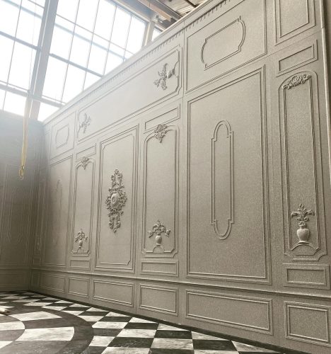 Große, verzierte Wand mit klassischem Stuckdekor und Reliefs neben einem Fenster, darunter ein Boden mit Schachbrettmuster.