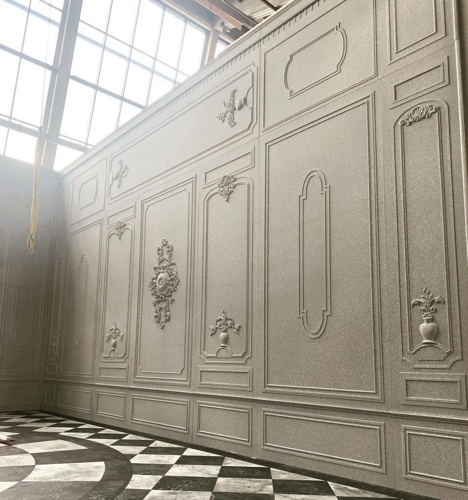 Große, verzierte Wand mit klassischem Stuckdekor und Reliefs neben einem Fenster, darunter ein Boden mit Schachbrettmuster.