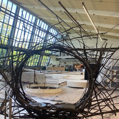 Große Halle mit hohen Fenstern, im Vordergrund eine kreisförmige Skulptur aus Ästen, im Hintergrund Werkstatt-Atmosphäre.