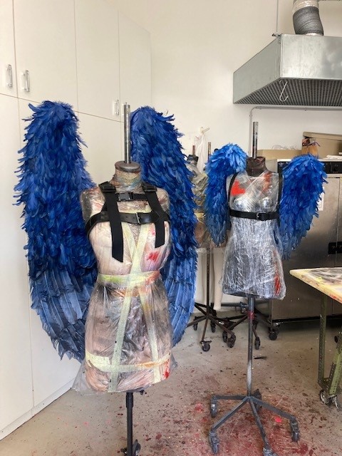 Zwei Schneiderpuppen mit blauen Engelsflügeln stehen in einem Atelier, umgeben von Werkstattmaterialien.