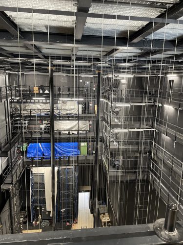Blick in den Bühnen- oder Technikbereich eines Theaters mit zahlreichen Seilzügen, Gerüsten und abgedeckten Ausstattungen.