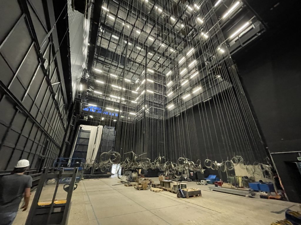 Blick hinter die Kulissen einer Theaterbühne mit vielen Seilzügen, Technikgeräten und einer Person mit Schutzhelm.