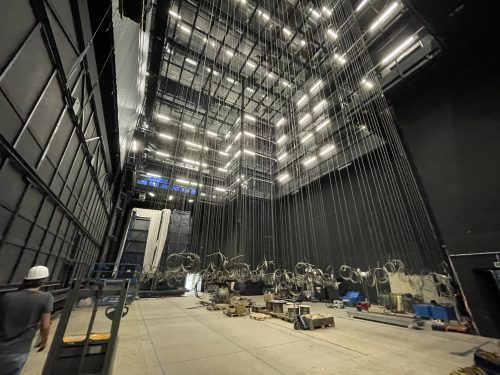 Blick hinter die Kulissen einer Theaterbühne mit vielen Seilzügen, Technikgeräten und einer Person mit Schutzhelm.