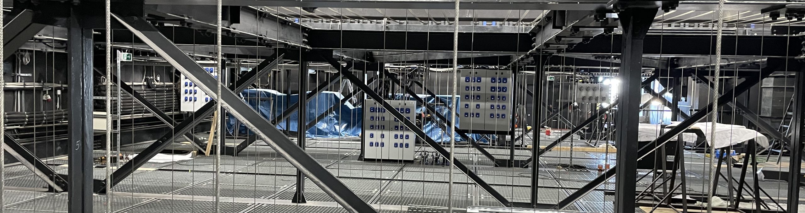 Industriehalle mit Metallgerüsten, Kabeln und mehreren weißen Schalttafeln im Hintergrund.