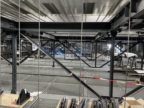 Stahlträger und Gitterroste in einer industriellen Halle mit technischen Anlagen und reflektierenden Oberflächen.