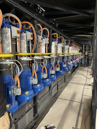 Eine Reihe von elektrischen Motoren mit blauen Gehäusen und orangenen Kabeln ist in einer technischen Anlage installiert.