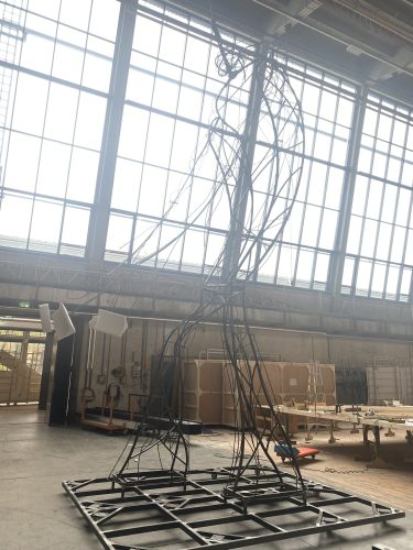 Hohe, abstrakte Skulptur aus gebogenen Metallstangen in einer Werkhalle mit großen Fenstern und Werkbänken im Hintergrund.