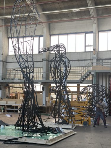 Zwei große Metallskulpturen in Vogelform stehen in einer Werkhalle, während eine Person an einer gelben Hebebühne arbeitet.