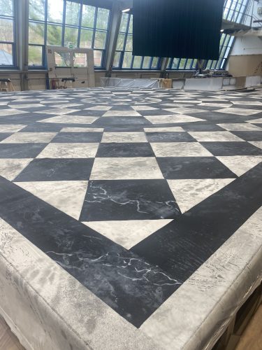 Großes, geometrisches Schachbrettmuster in Schwarz und Weiß auf einer ebenen Fläche in einem hellen Raum mit großen Fenstern.