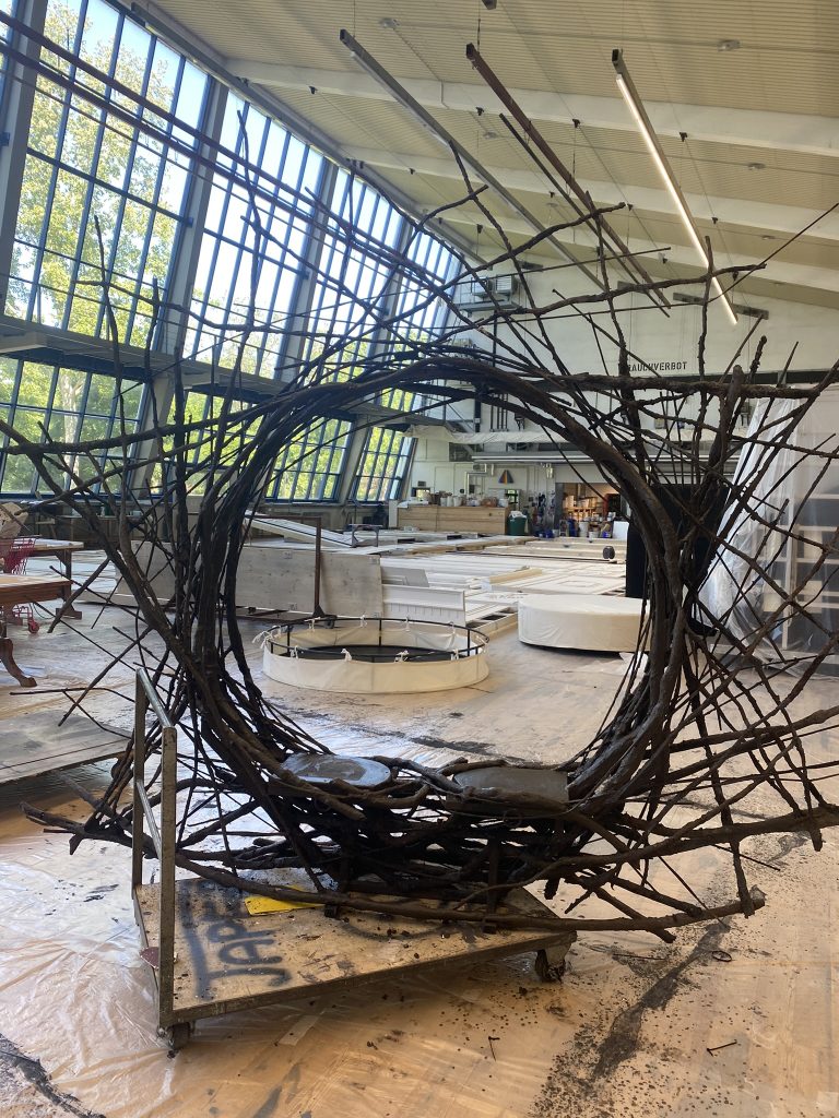Großes, kunstvoll geformtes Nest aus verschlungenen Ästen steht in einer Werkstatt mit hohen Fenstern und Holzboden.