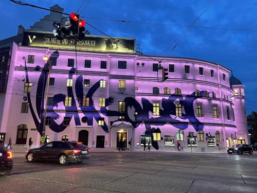 Ein großes, hell beleuchtetes Gebäude mit lila Fassadenbeleuchtung und auffälligem Graffiti sowie vorbeifahrenden Autos bei Abenddämmerung.