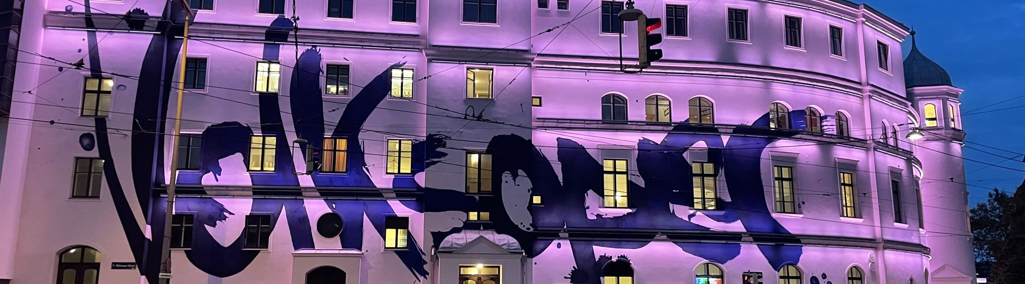 Großes weißes Gebäude mit lilafarbener Beleuchtung und einer großen, schwarzen Graffiti-Schrift an der Fassade.