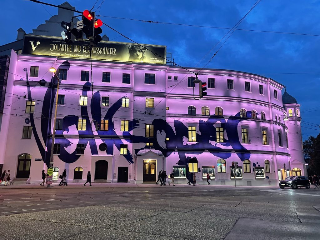 Ein großes Gebäude mit lila Beleuchtung und großflächigem Graffiti-Schriftzug „Volksoper“ an der Fassade bei Abenddämmerung.