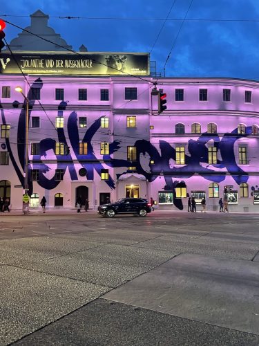 Ein Gebäude mit lila Beleuchtung und großem Graffiti, davor fährt ein schwarzes Auto bei Abenddämmerung vorbei.