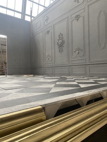 Eine erhöhte Bühne mit schwarz-weiß gemustertem Boden und einer grauen Wand im klassischen Stil mit Verzierungen im Hintergrund.